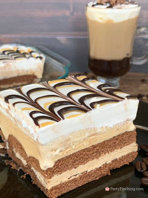 A square slice of layered cake with a chocolate marbled topping sits next to a glass of coffee on a dark surface.