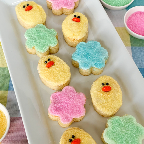 Mini pound cakes shaped like chicks and flowers, decorated with colorful sanding sugar, on a long white serving platter.