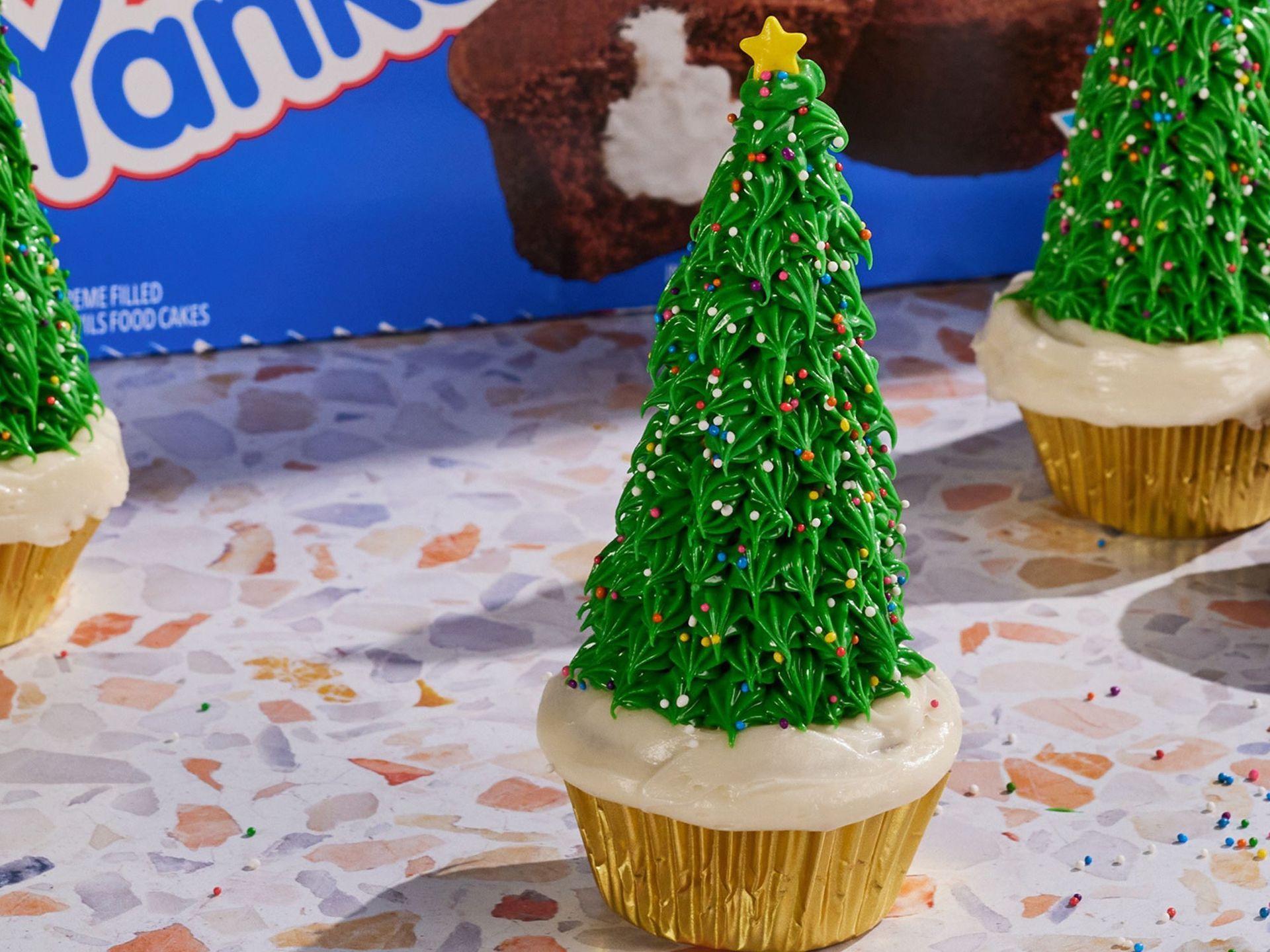 Three cupcakes decorated to look like Christmas trees with green piped icing and stars on a speckled surface.