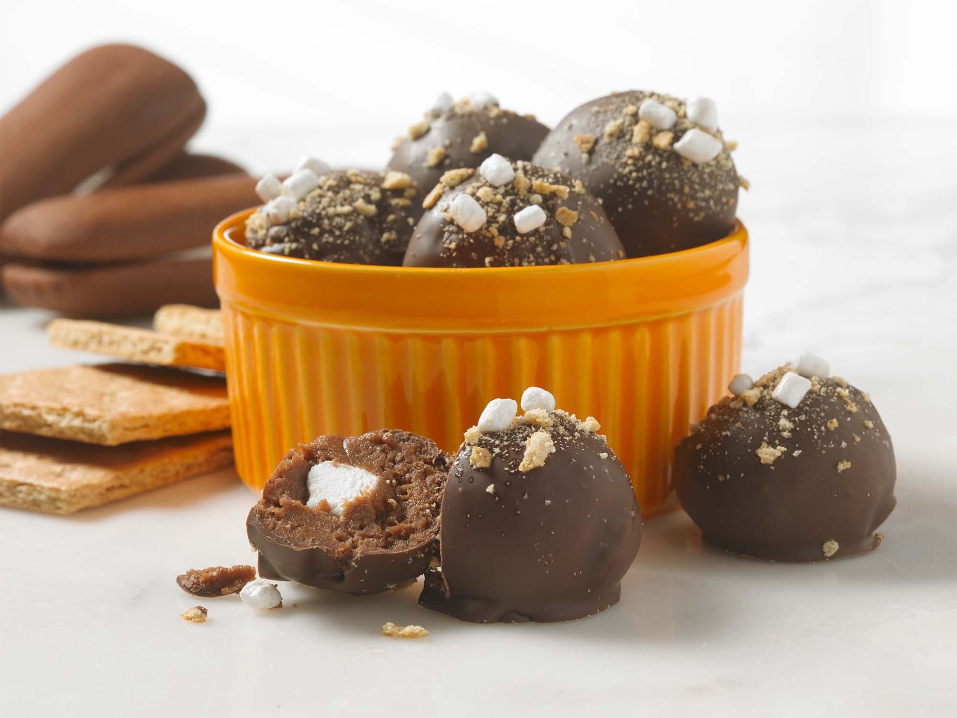 Chocolate s’mores truffles topped with marshmallow bits and graham cracker crumbs in an orange ramekin on a white surface.