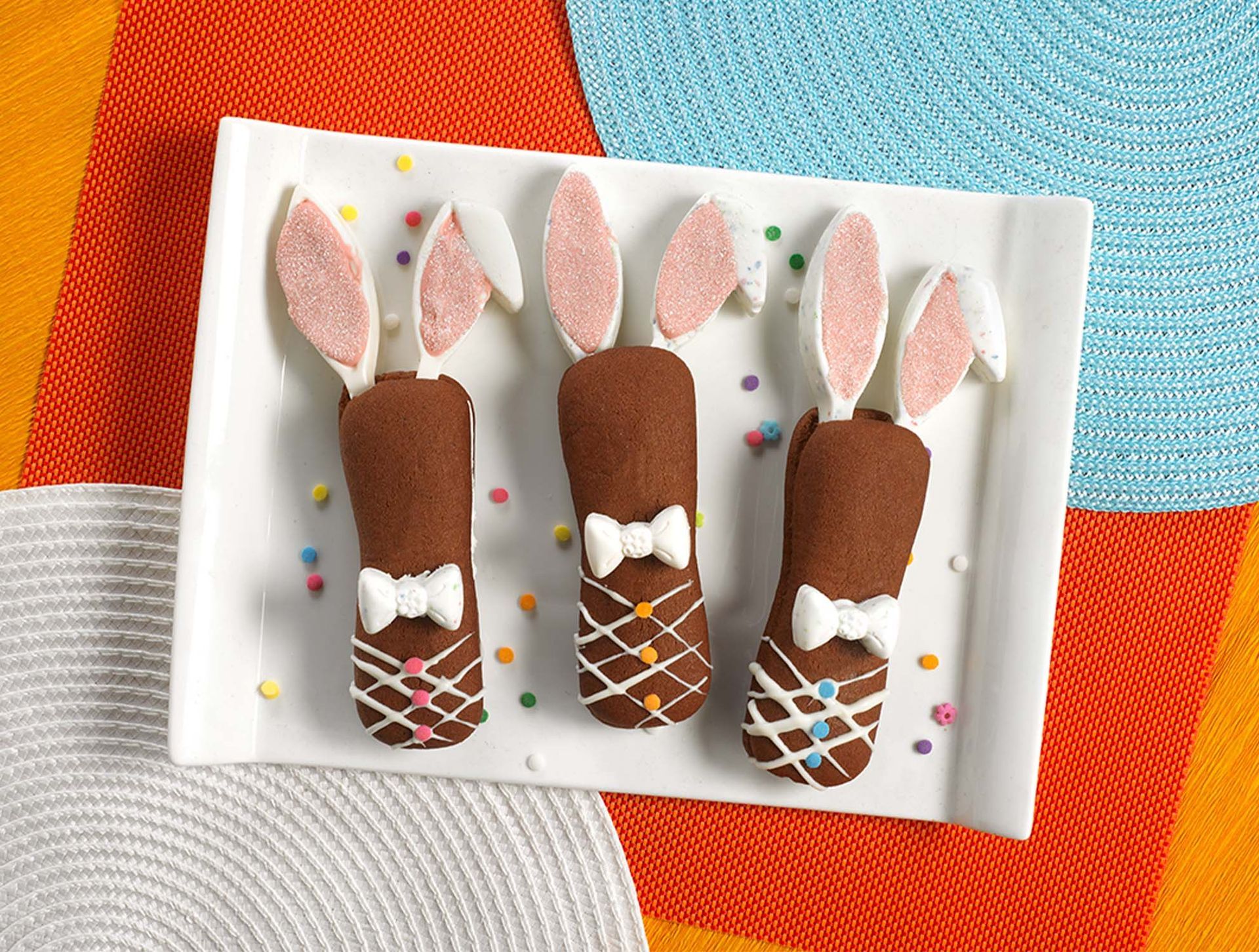 Three brown chocolate-coated treats decorated as Easter bunnies with marshmallow ears and icing ties on a white plate.