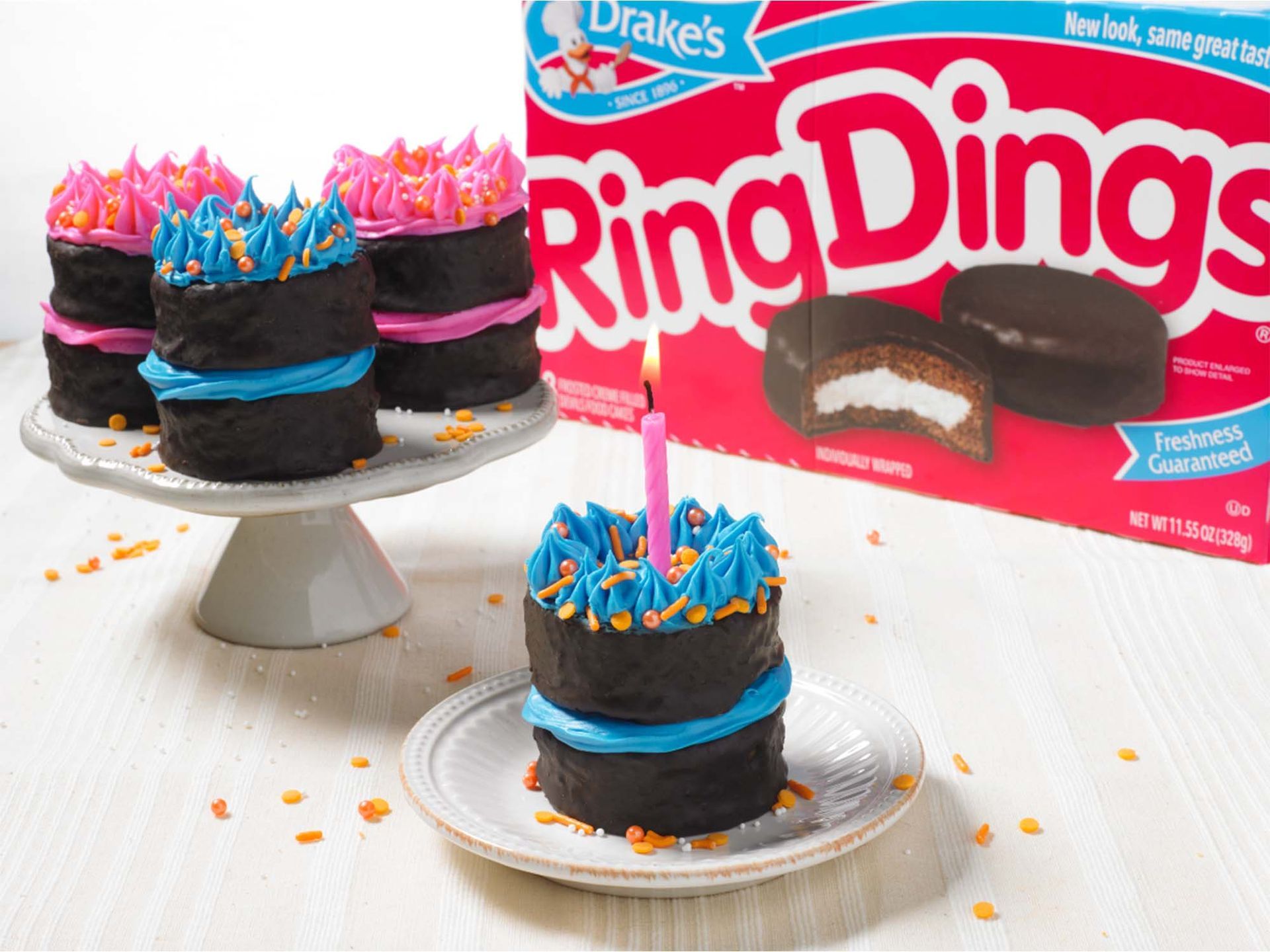 Three mini chocolate cakes decorated with pink or blue icing sit on white plates, with a Ring Ding box in the background.