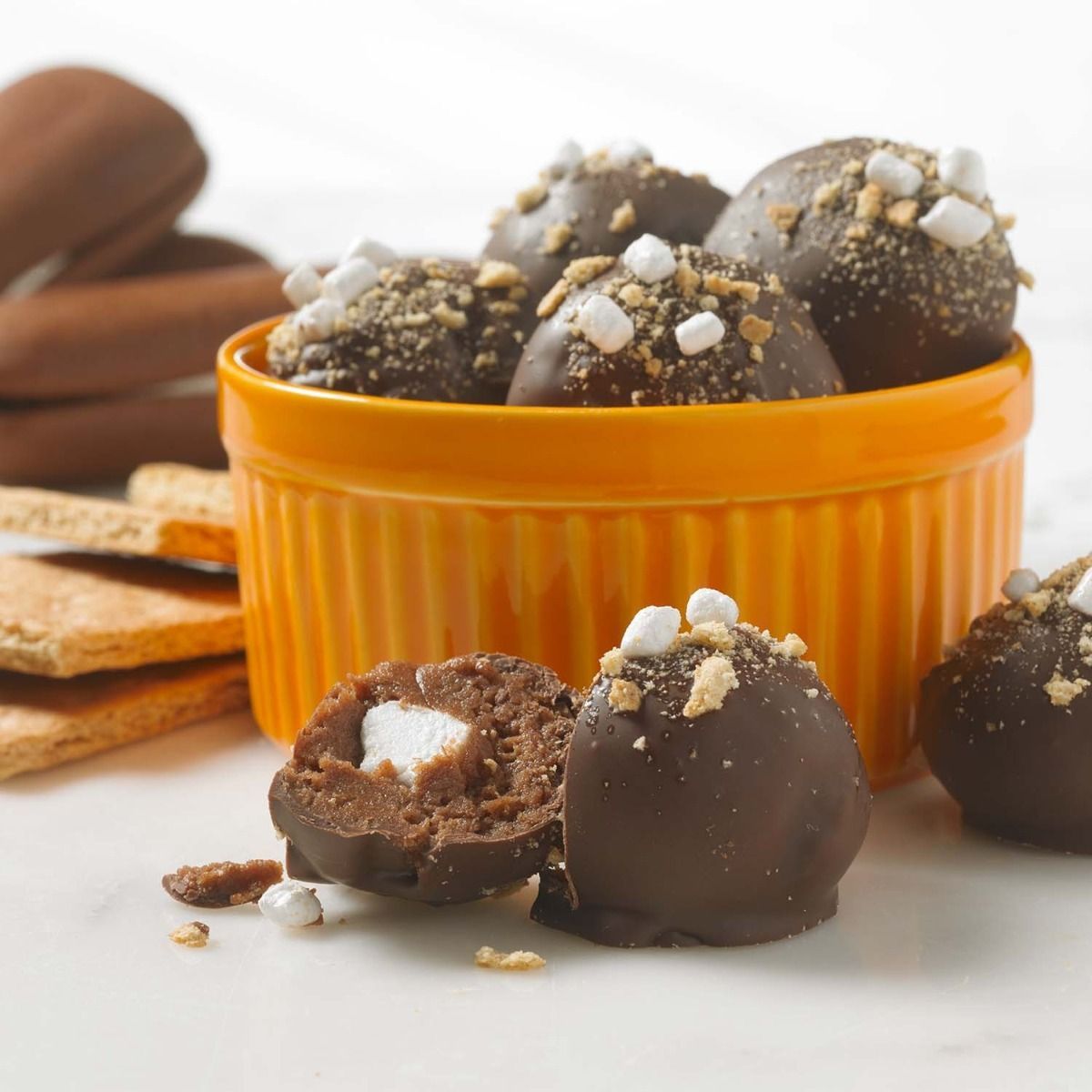 Chocolate-covered s'mores truffles with marshmallow bits in an orange bowl, with graham crackers on a white surface.
