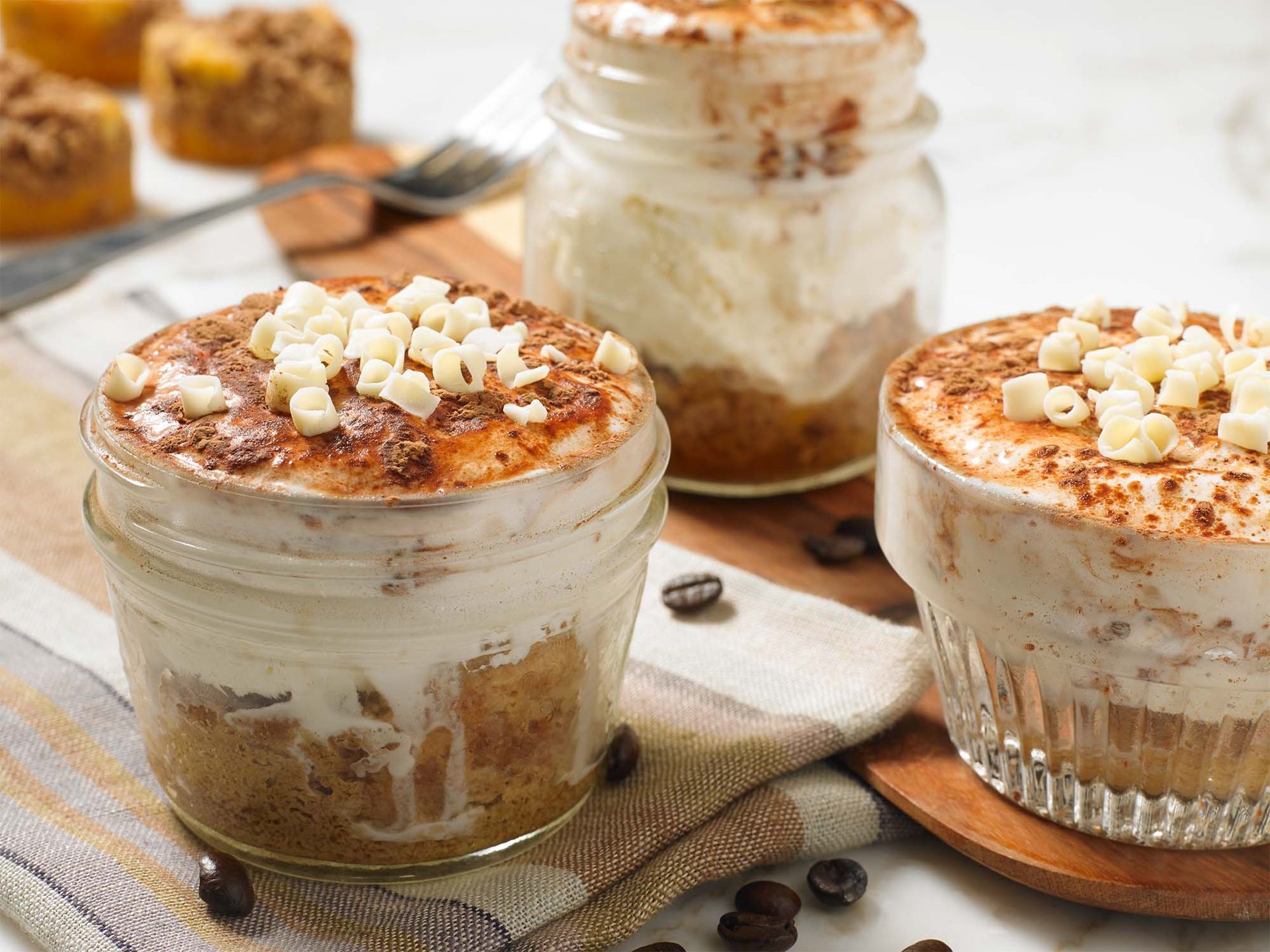 Three glass jars filled with layered tiramisu, topped with cream, cocoa powder, and white chocolate shavings.