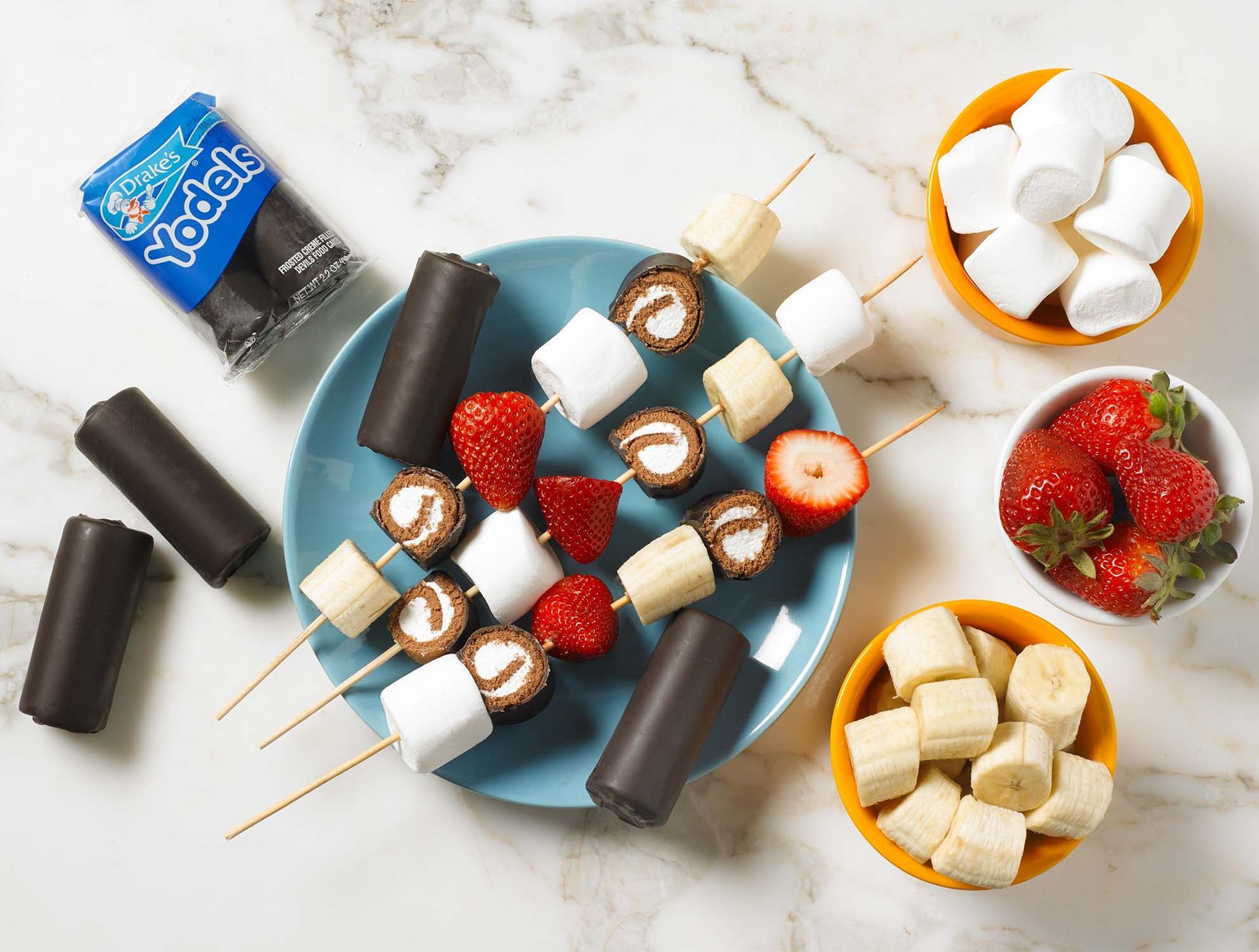 A blue plate with fruit and Yodels skewers, surrounded by bowls of marshmallows, strawberries, and banana slices.