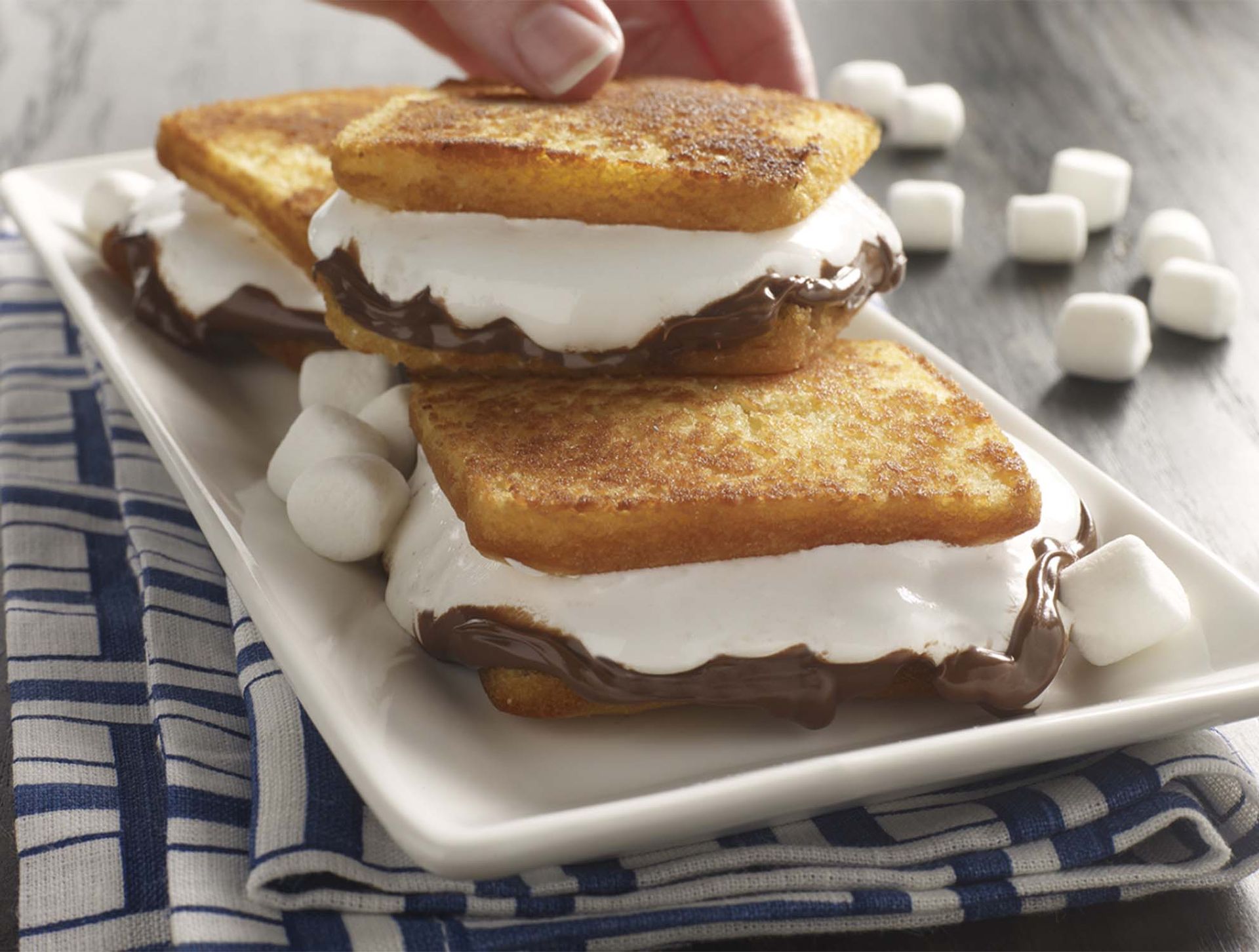 Two French toast sandwiches filled with melted chocolate and marshmallow on a white serving platter, surrounded by marshmallows.