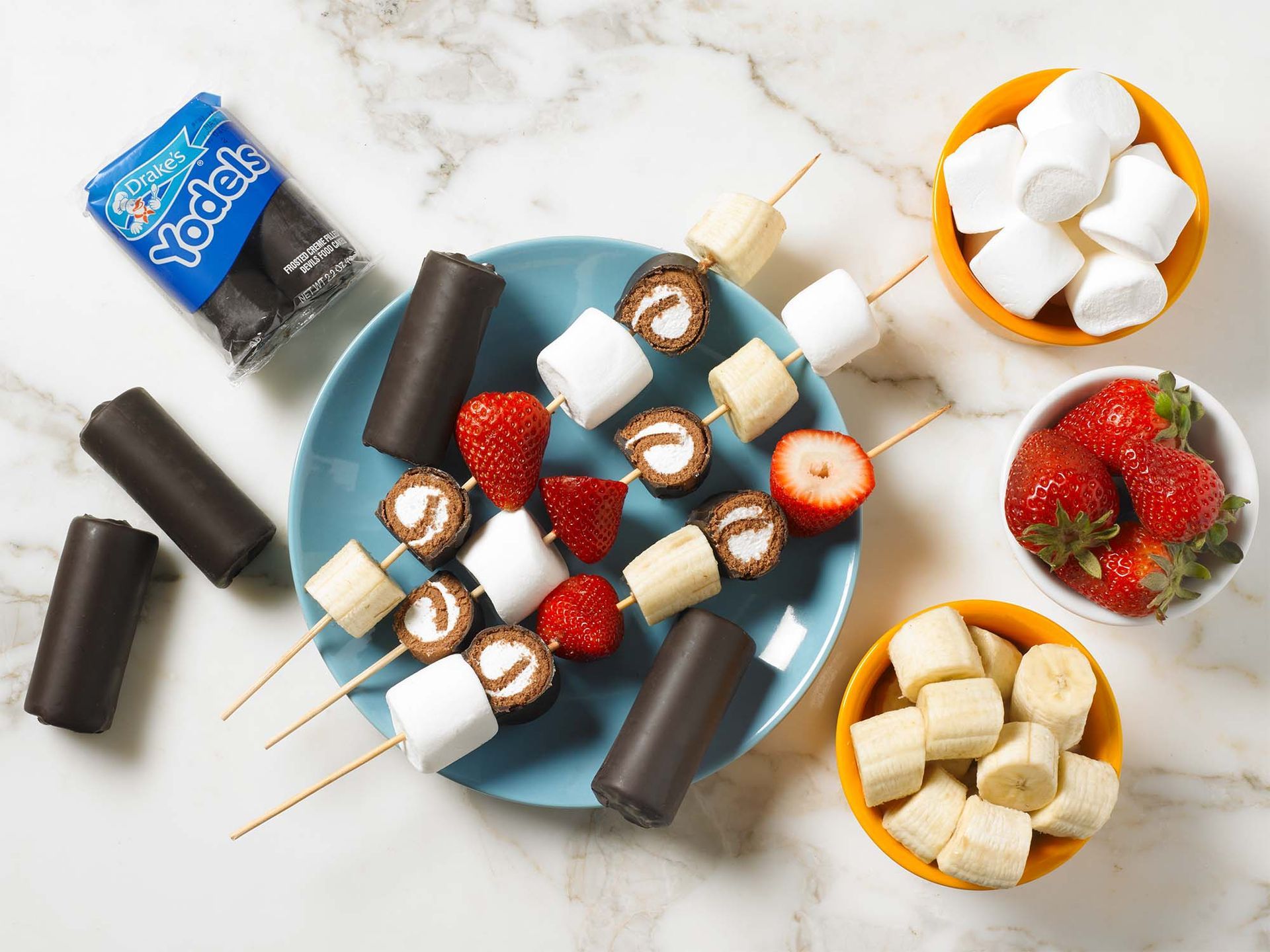 Three skewers with Yodels, marshmallows, strawberries, and bananas on a blue plate, with bowls of the ingredients nearby.