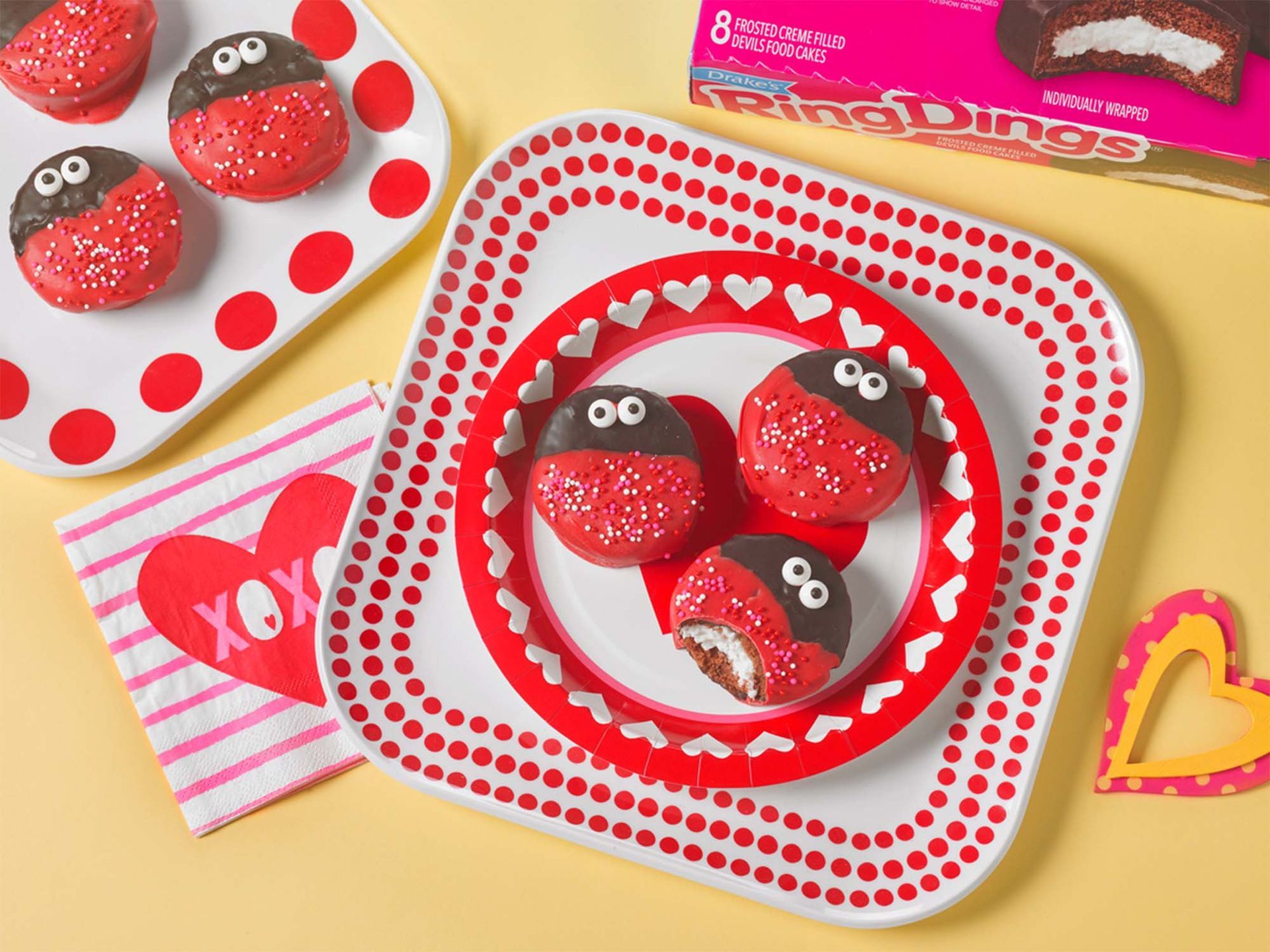 Ladybug-decorated Ring Dings on patterned plates and a napkin with a heart, set against a yellow background.