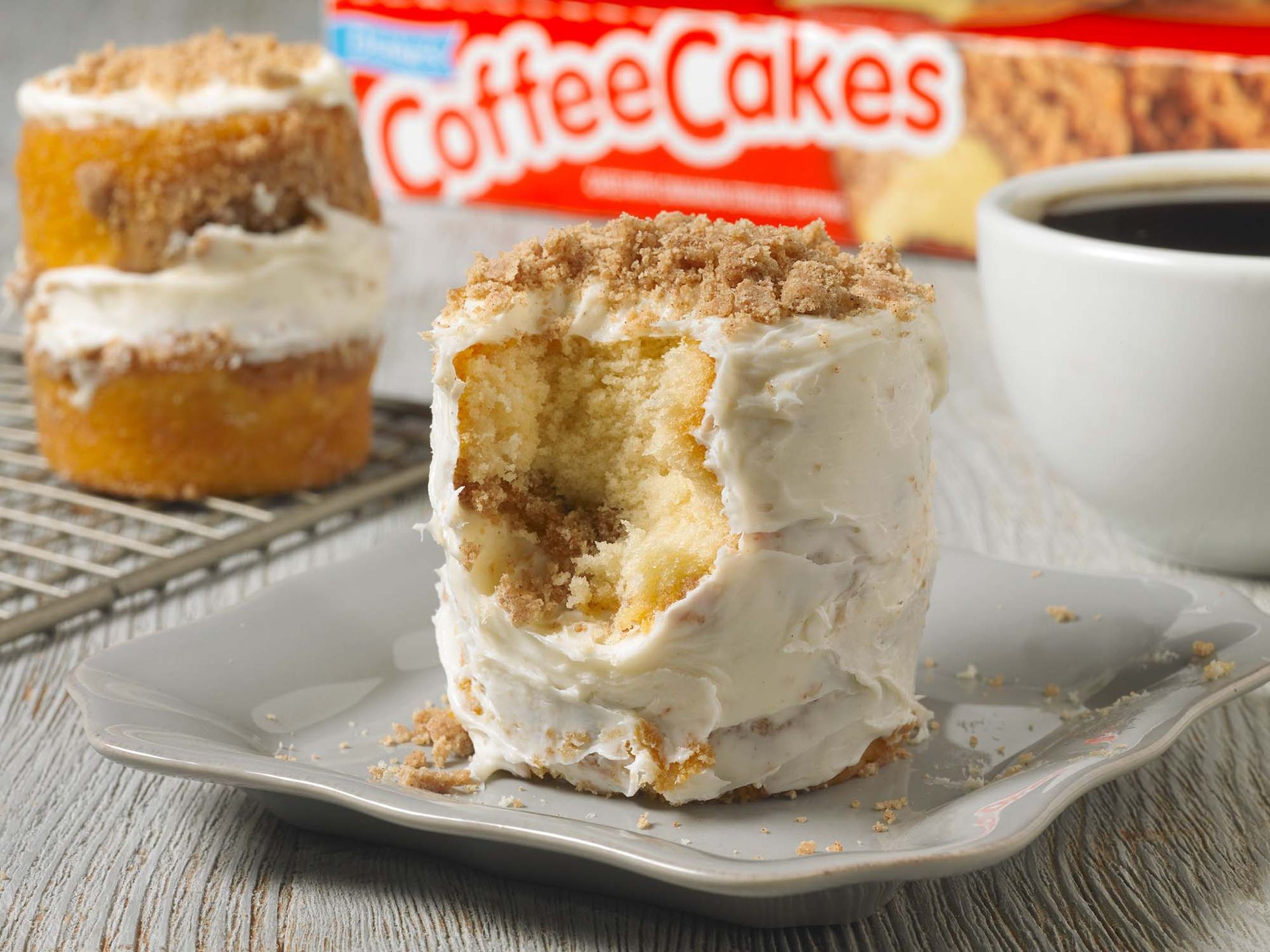 Two small, crumb-topped cakes with frosting sit on a table next to a cup of coffee and a box of coffee cakes.
