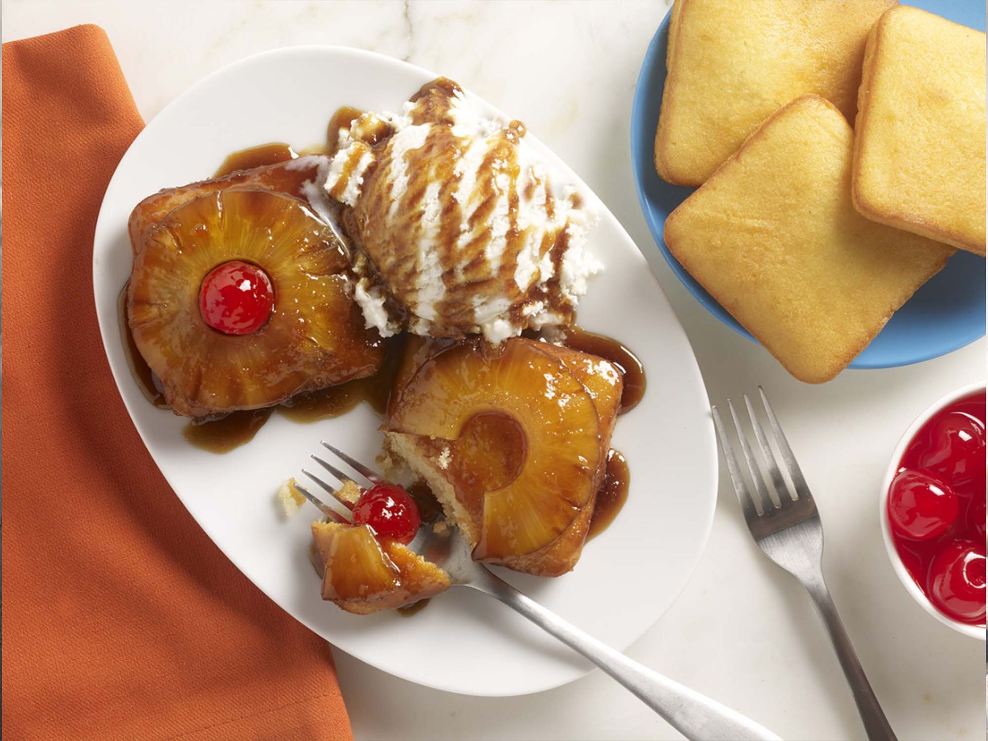 Two pineapple upside-down cakes with cherries and whipped cream on a white plate, alongside extra cakes and a fork.