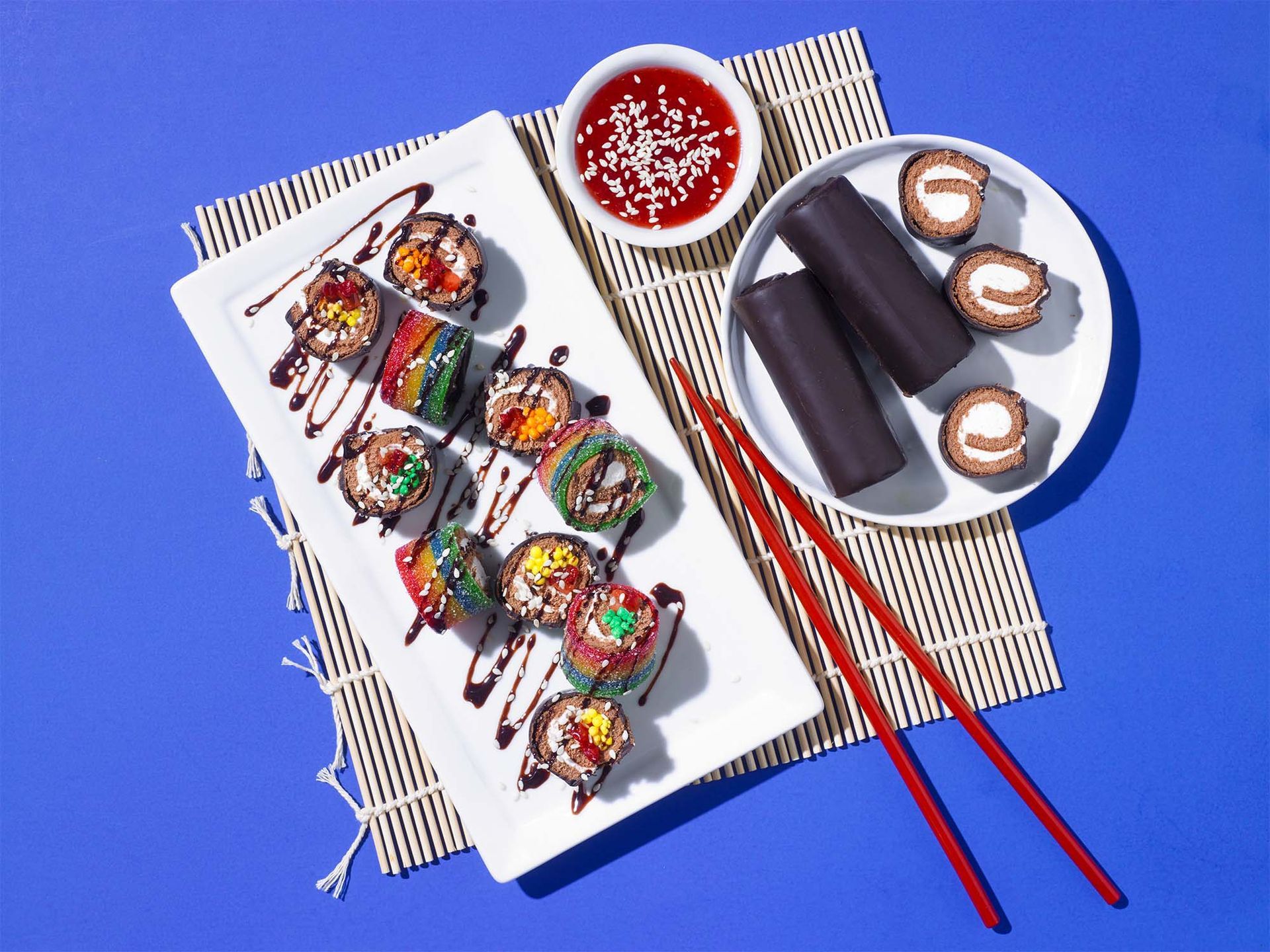 Dessert sushi rolls, chocolate-covered treats, and red dipping sauce on a bamboo mat with red chopsticks against blue.