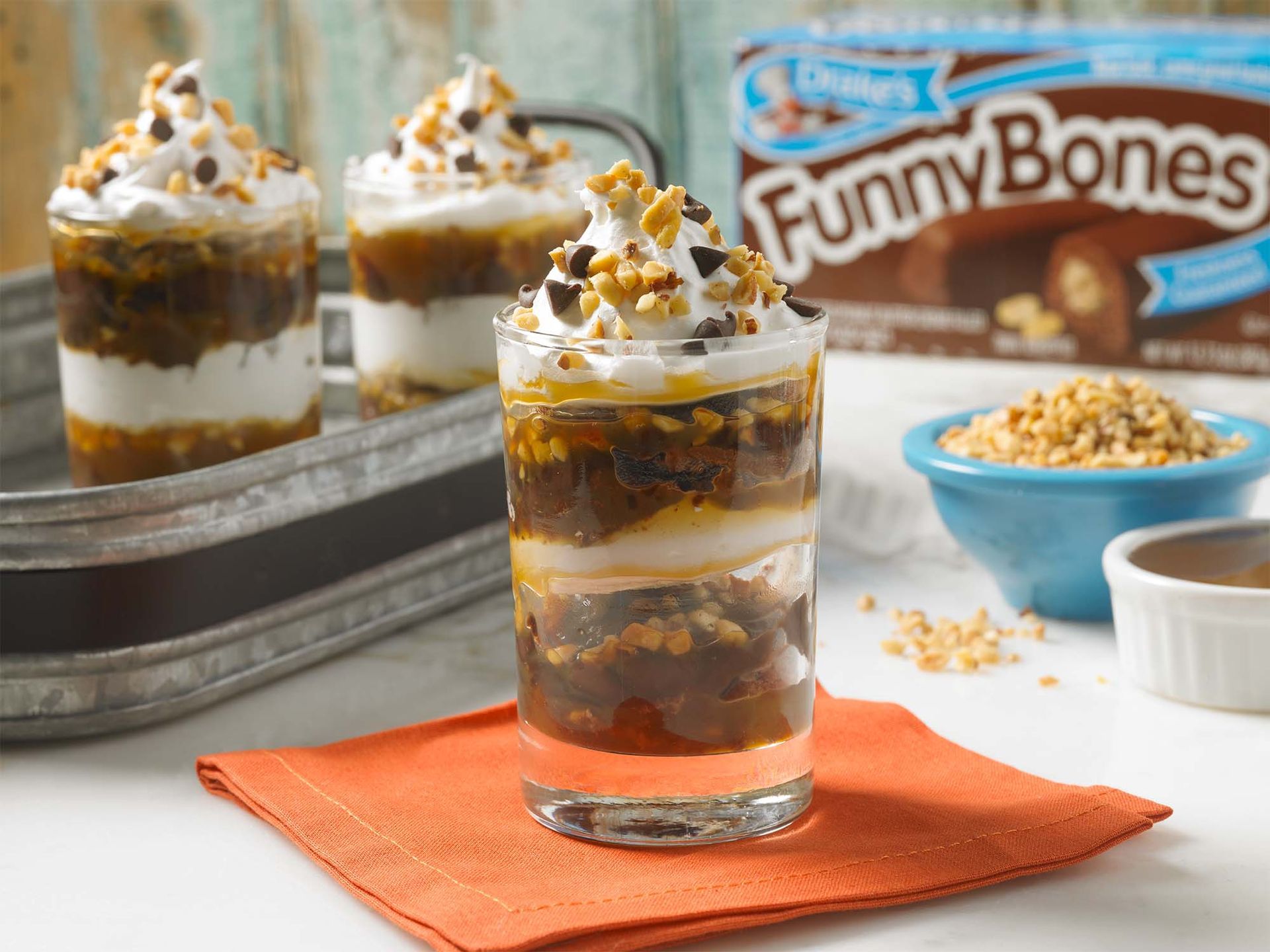 Three parfaits made with Funny Bones, whipped cream, and nuts sit on a rustic tray and an orange napkin.
