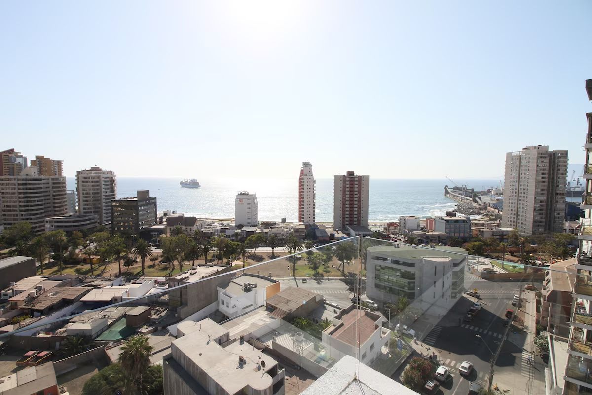 Paisaje urbano con vistas al océano, con edificios, un parque y un barco bajo un cielo azul claro.