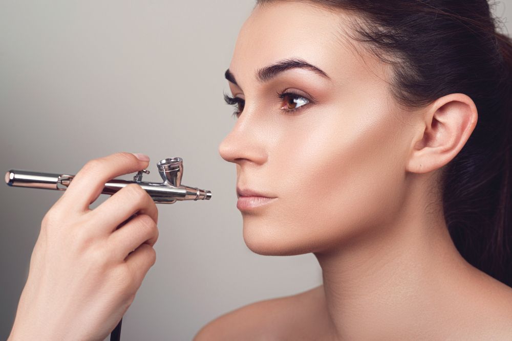 Woman applying makeup with an airbrush tool, close-up profile against a plain background
