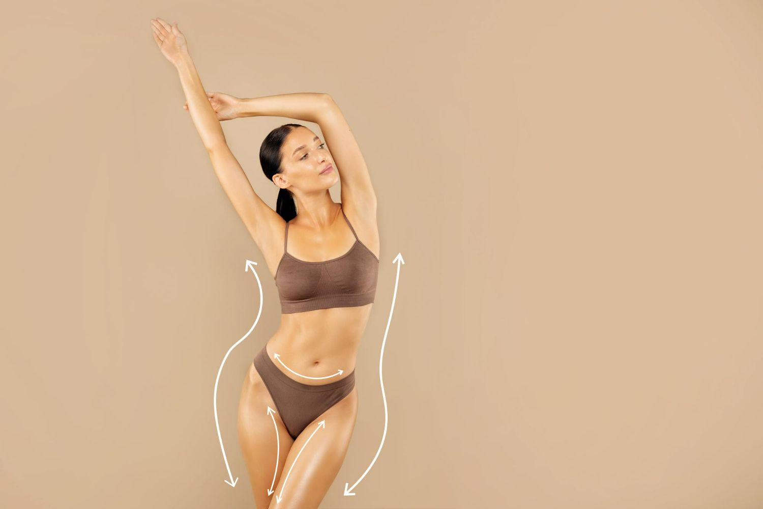 Woman in brown lingerie posing against a beige backdrop with arms raised overhead.