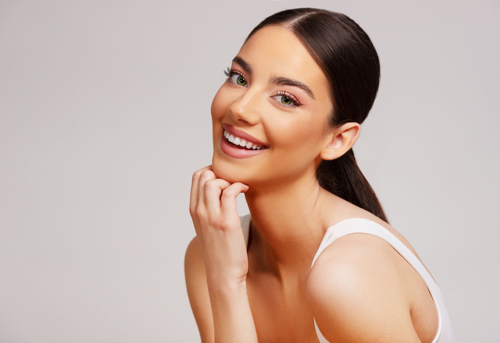 A woman in a white tank top is smiling with her hand on her chin.