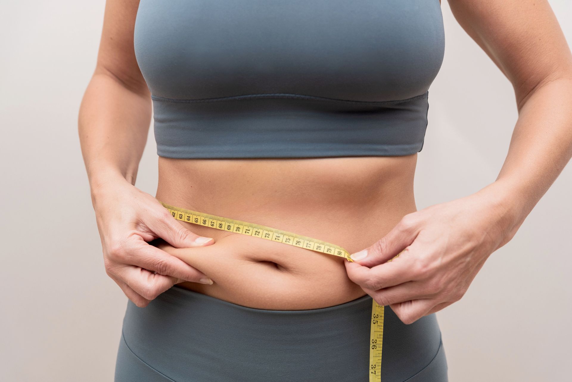 Person measuring waist with a yellow tape measure against a light gray background.