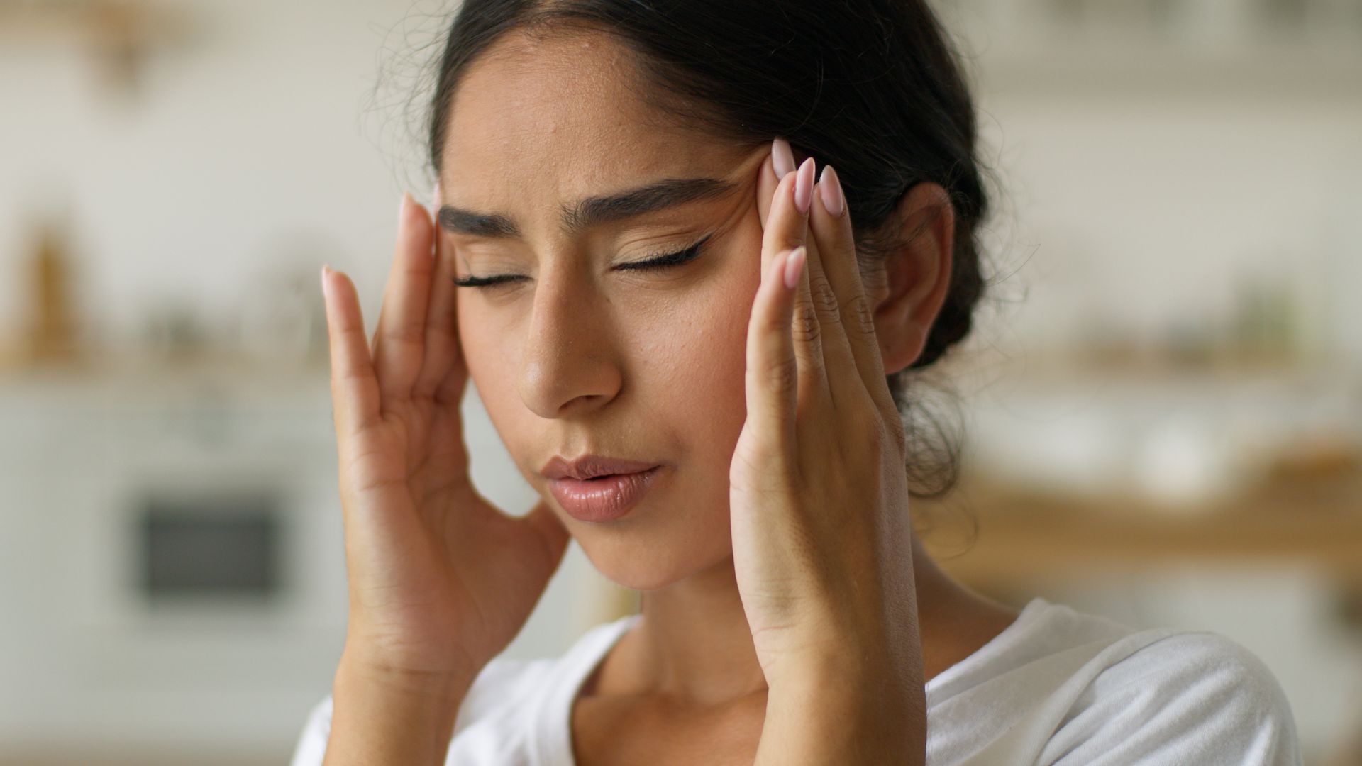 Woman holding her temples with eyes closed, appearing to have a headache in a kitchen.