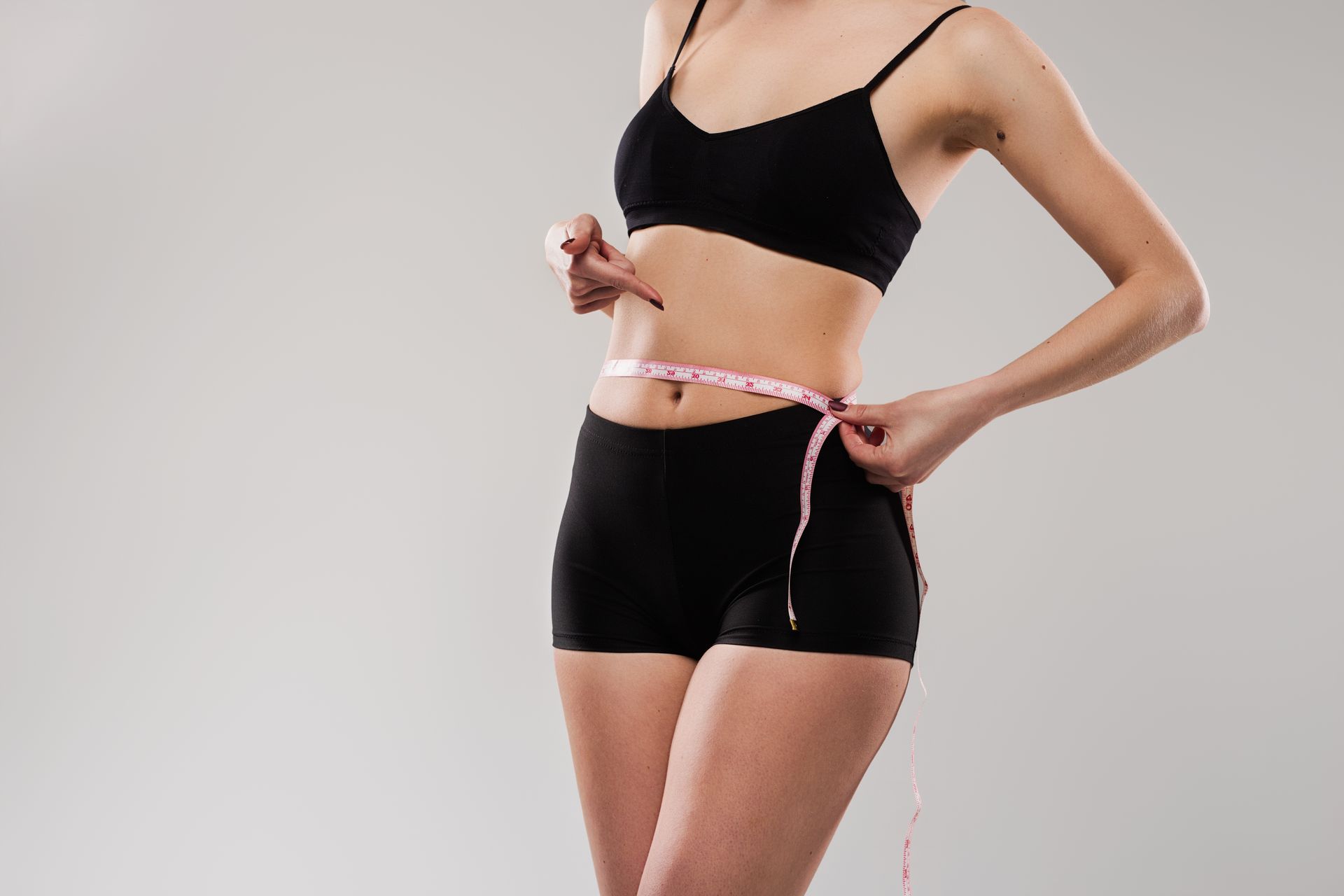 Person in black sports bra and shorts measuring waist with a pink tape measure.