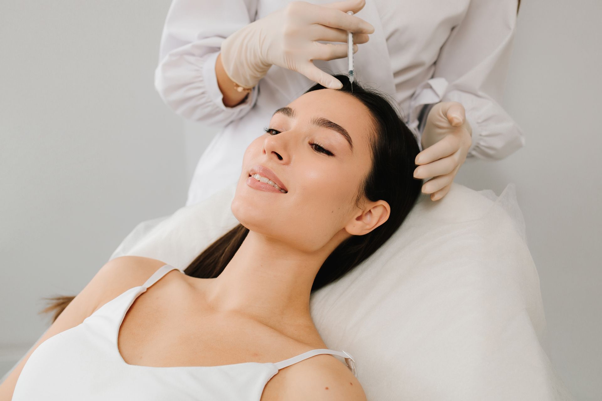 Woman receiving a facial treatment while lying on a treatment bed, with a technician applying a serum dropper
