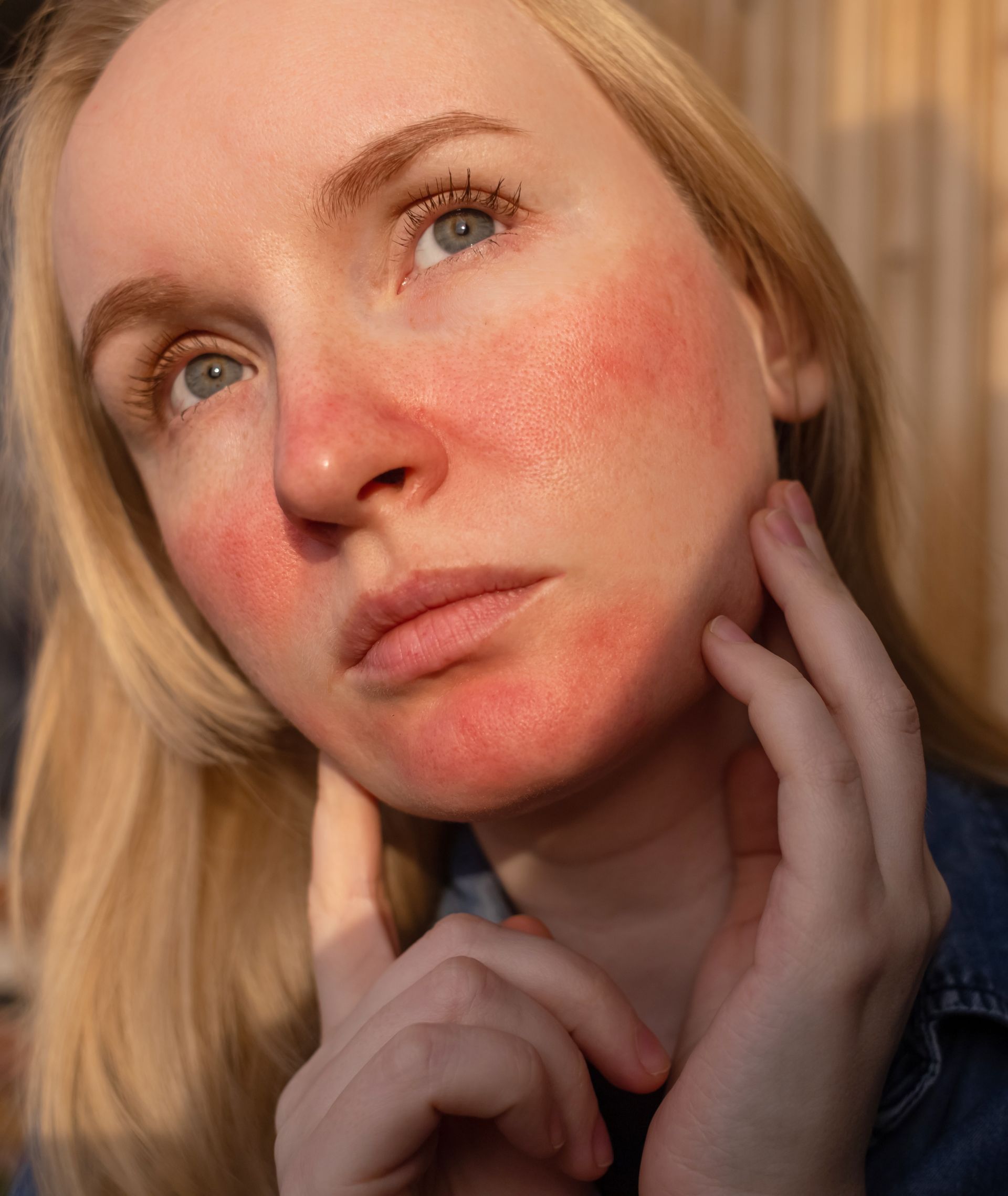 Close-up portrait of a person with flushed cheeks, looking upward and touching their face in warm light