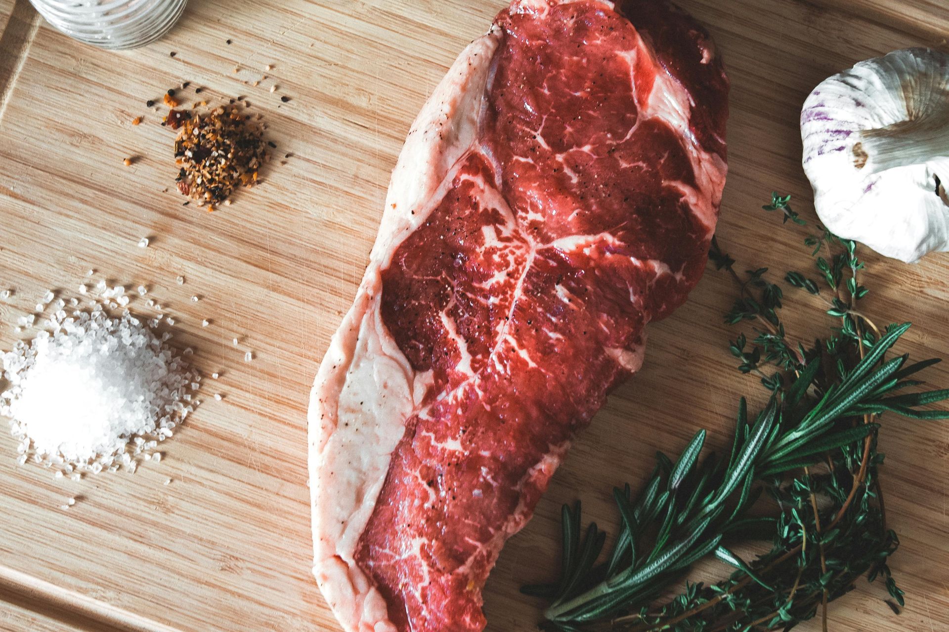 Raw steak on a wooden cutting board with salt, pepper, garlic, and rosemary herbs