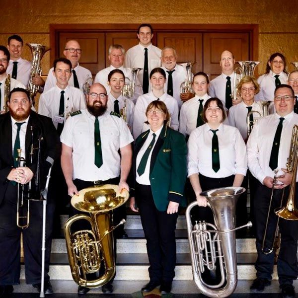 A brass band ensemble in white shirts and green ties posing together indoors on a set of steps, some holding instruments.