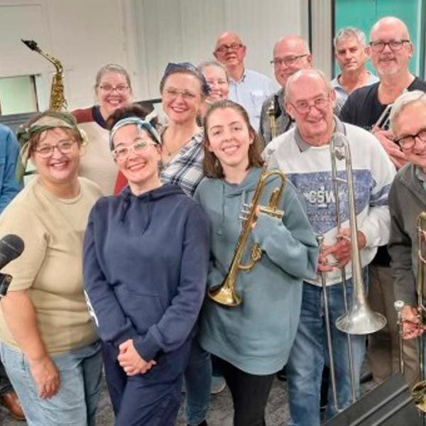 A group of people standing together in a room, some holding brass musical instruments like trumpets and a trombone.