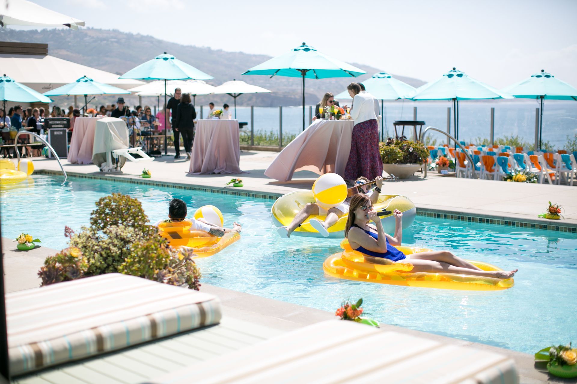 People relaxing on yellow inflatables in a sunny outdoor pool surrounded by tables and umbrellas.