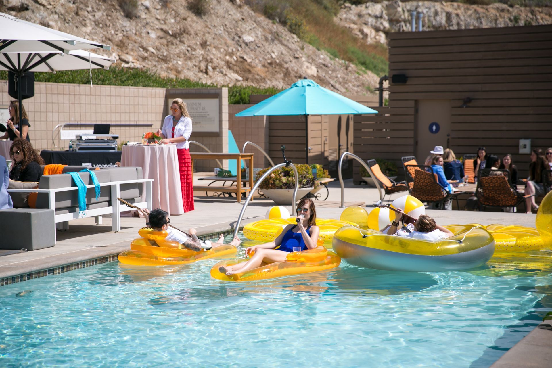 People relax on yellow inflatable rafts in a resort pool near a hillside, while others socialize on the nearby deck.