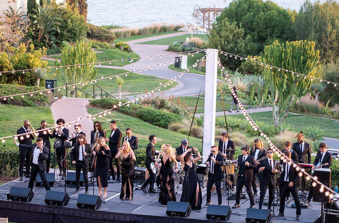 A band in black formal wear performs on an outdoor stage at dusk, set against a green park, walking paths, and a water view.
