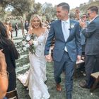 A newlywed couple walks hand-in-hand down an aisle outdoors, both smiling, surrounded by guests in a garden setting.