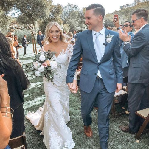 A newlywed couple walks hand-in-hand down an aisle outdoors, both smiling, surrounded by guests in a garden setting.