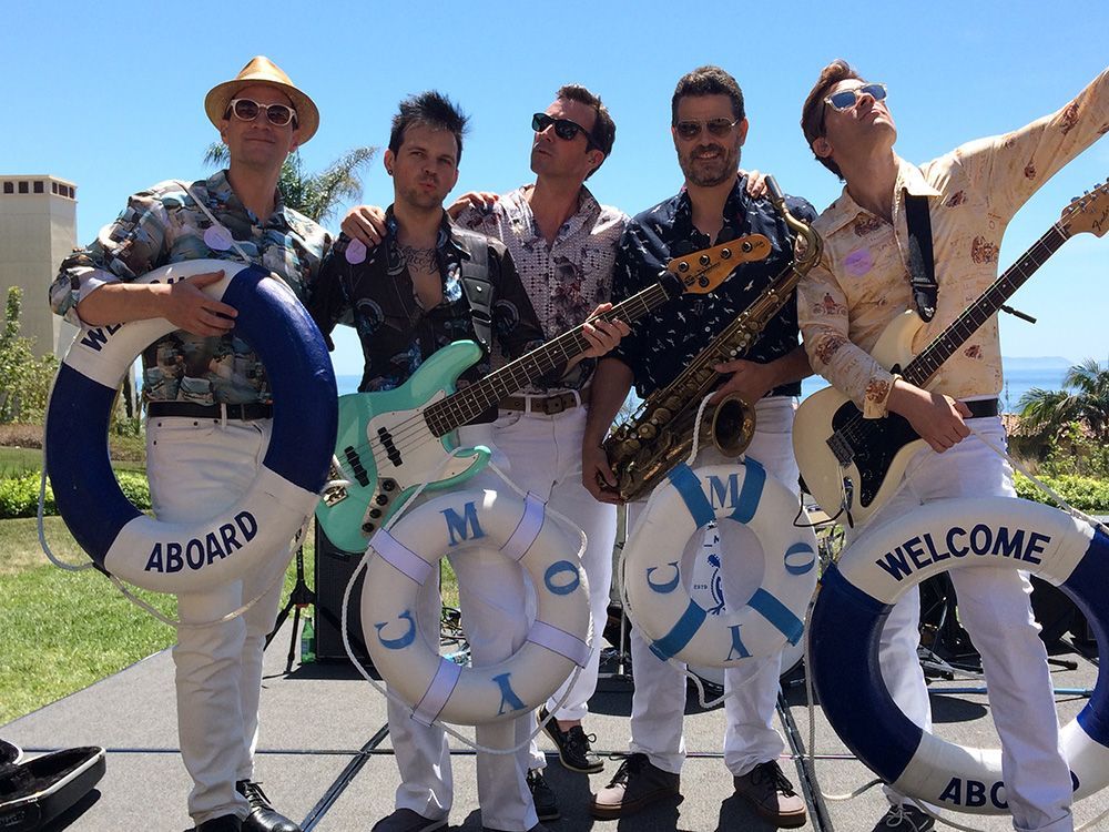 Five band members in light pants pose outdoors with instruments and 