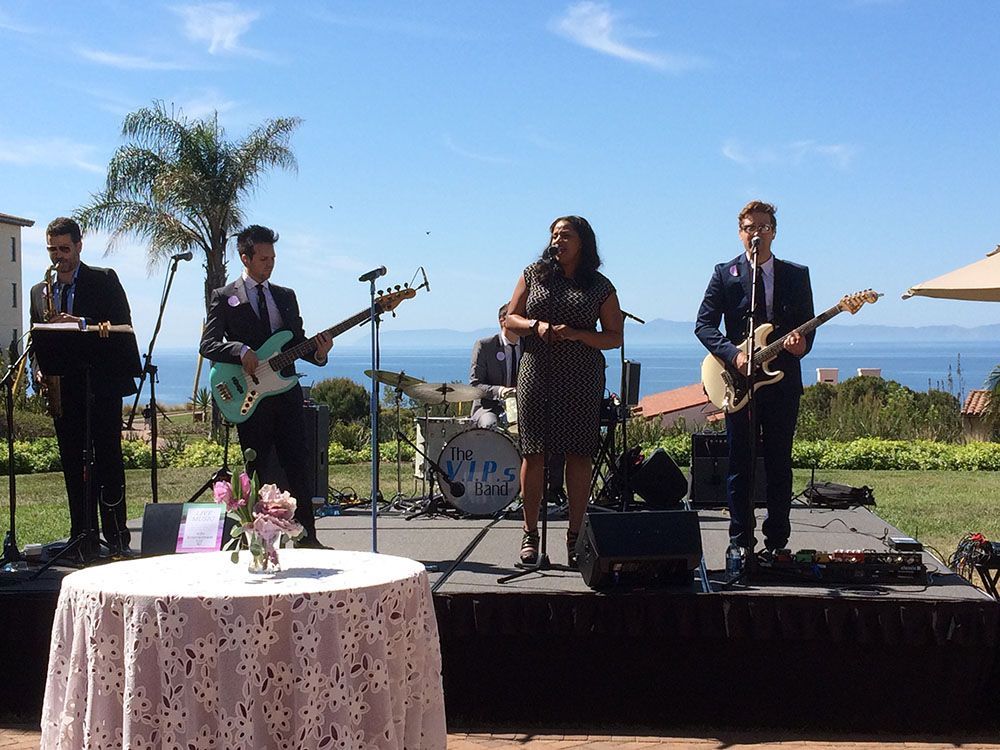 A band performing on an outdoor stage with ocean views, featuring a vocalist, two guitarists, and a drummer.