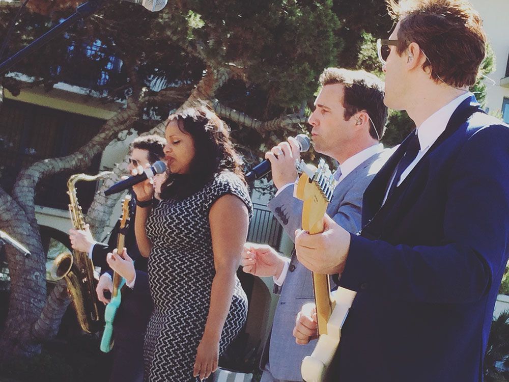 A band performing outdoors, with a singer, guitarist, saxophonist, and bassist, all dressed in formal attire.