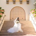 A bride in a white wedding gown holding a bouquet stands on patterned stairs in front of a decorative arched wall.