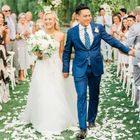 A bride and groom walk hand-in-hand down a flower-petal-strewn aisle, smiling as wedding guests look on.