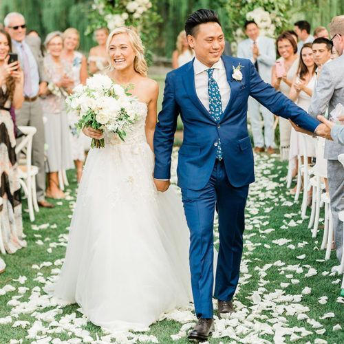 A bride and groom walk hand-in-hand down a flower-petal-strewn aisle, smiling as wedding guests look on.