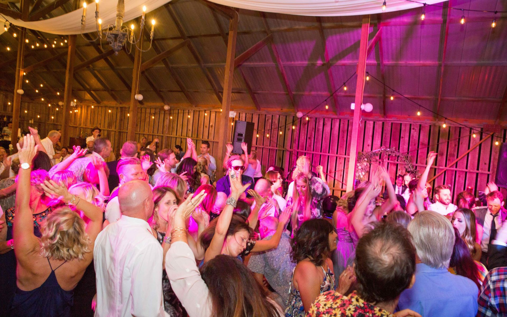 A crowd of people dances inside a rustic barn decorated with string lights and purple ambient lighting.