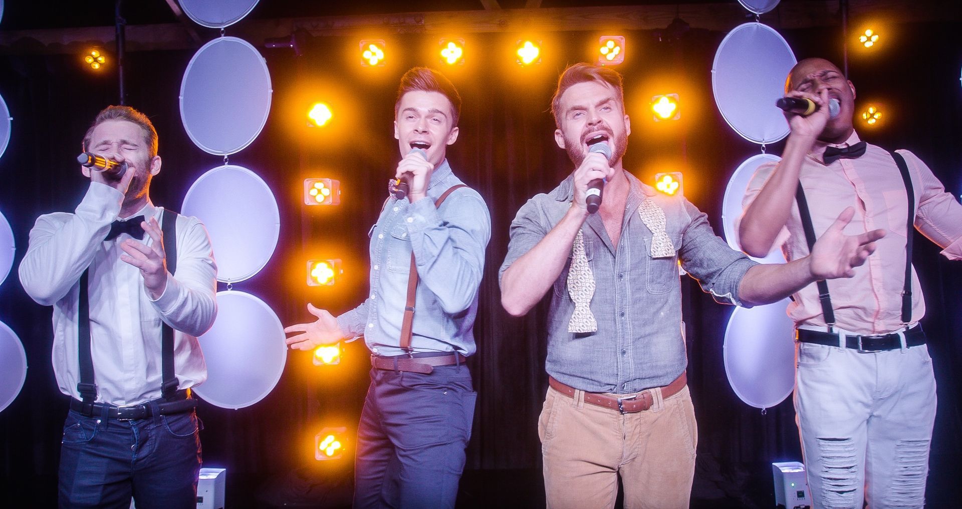 Four singers in button-down shirts and suspenders perform into microphones on a stage with glowing circular lights.