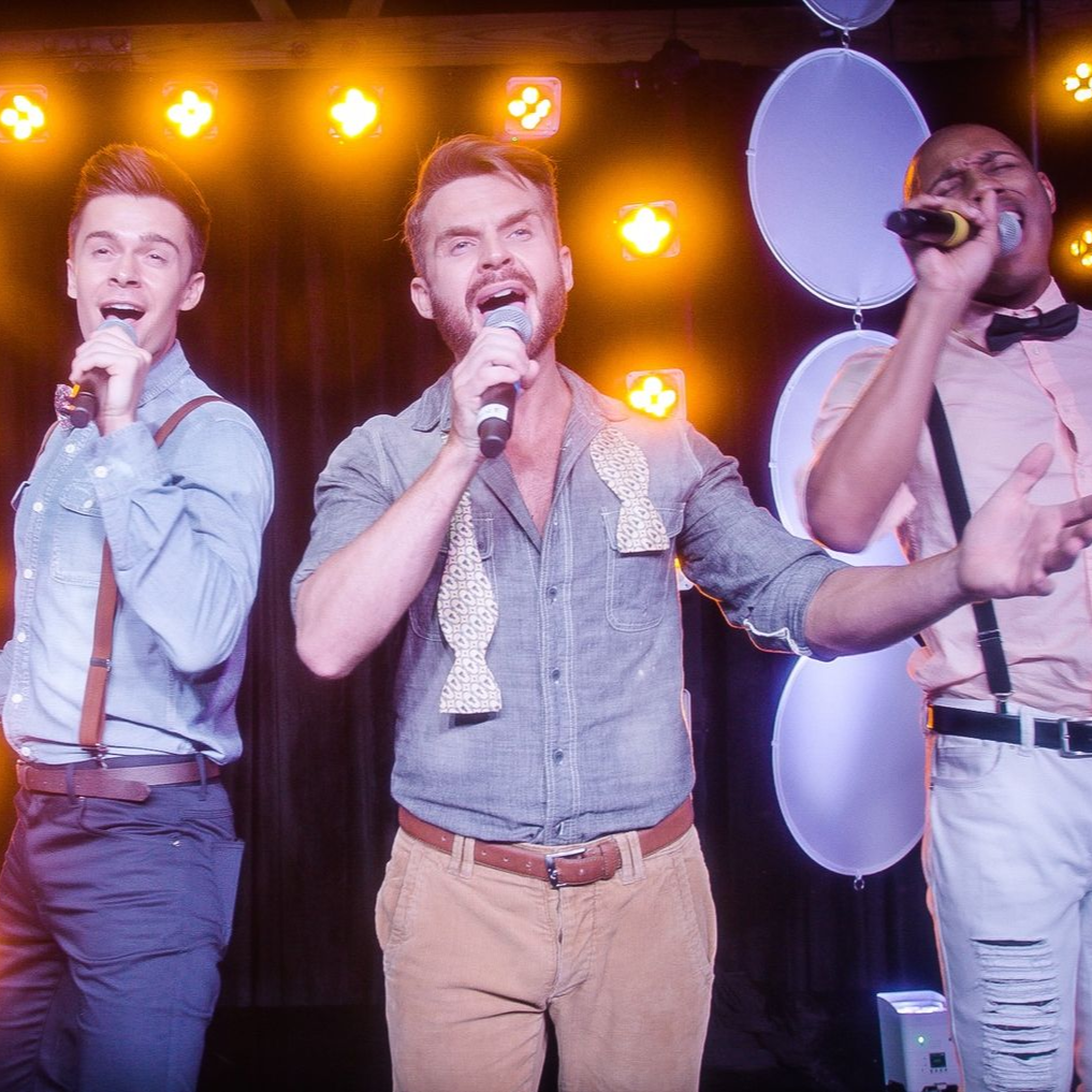 Three performers sing into microphones on a stage illuminated by warm, circular lights.