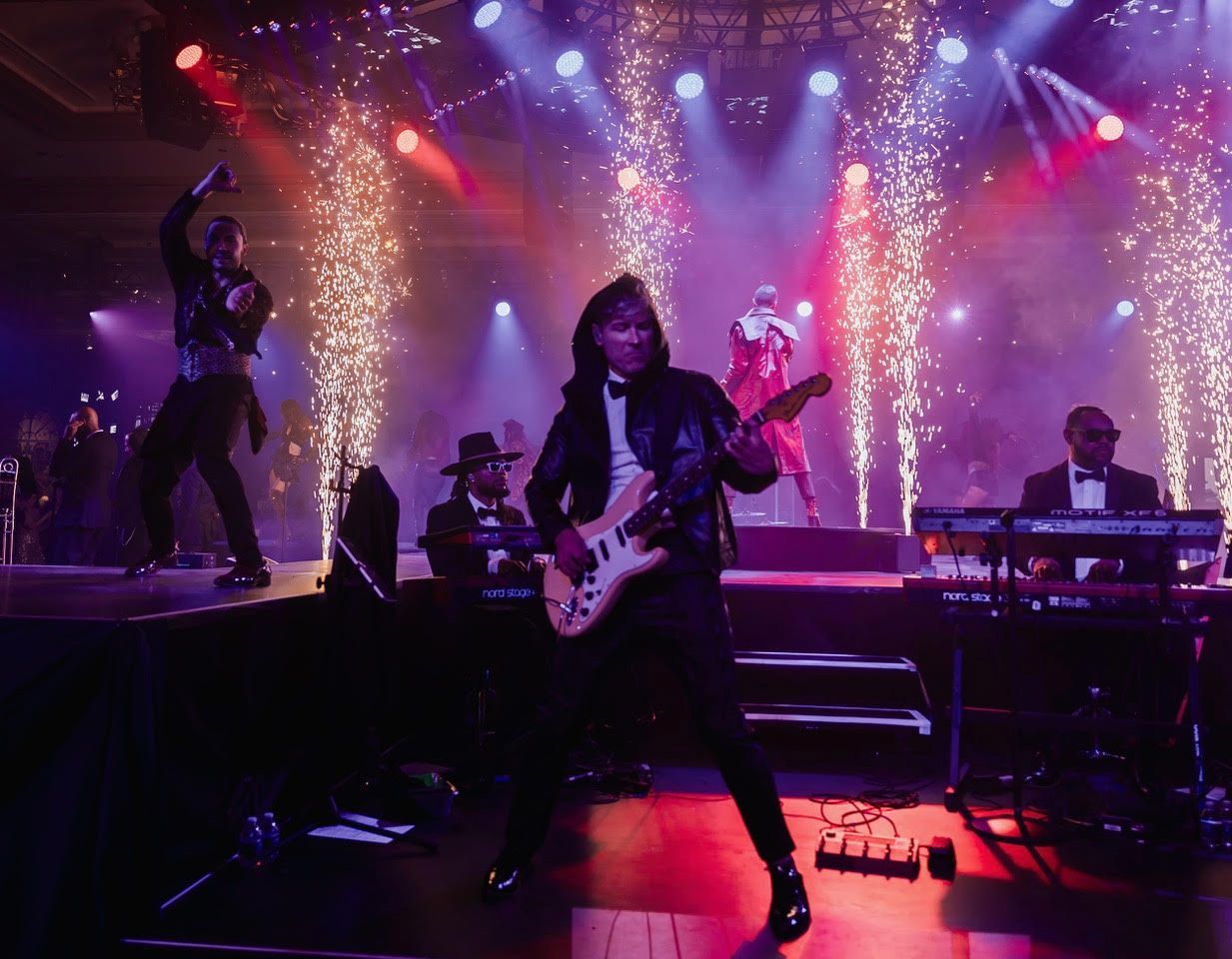 A band performs on a stage lit by spotlights and pyrotechnics, featuring a guitarist in the center and a keyboardist.