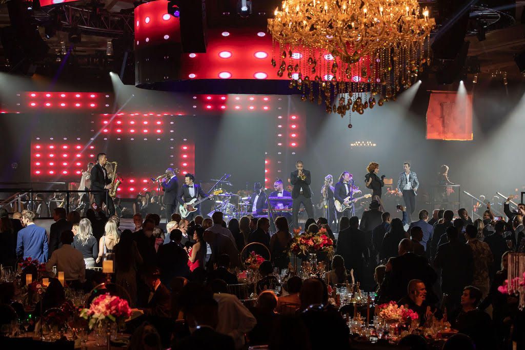 A band performs on a stage with red light panels and a chandelier at a dimly lit, crowded formal event.