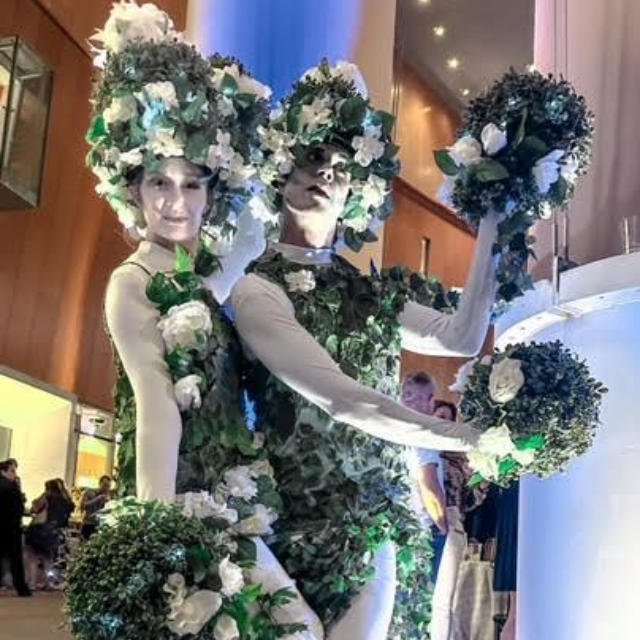 Two performers in floral, vine-covered costumes and white face paint pose, holding round bouquets, at an evening event.