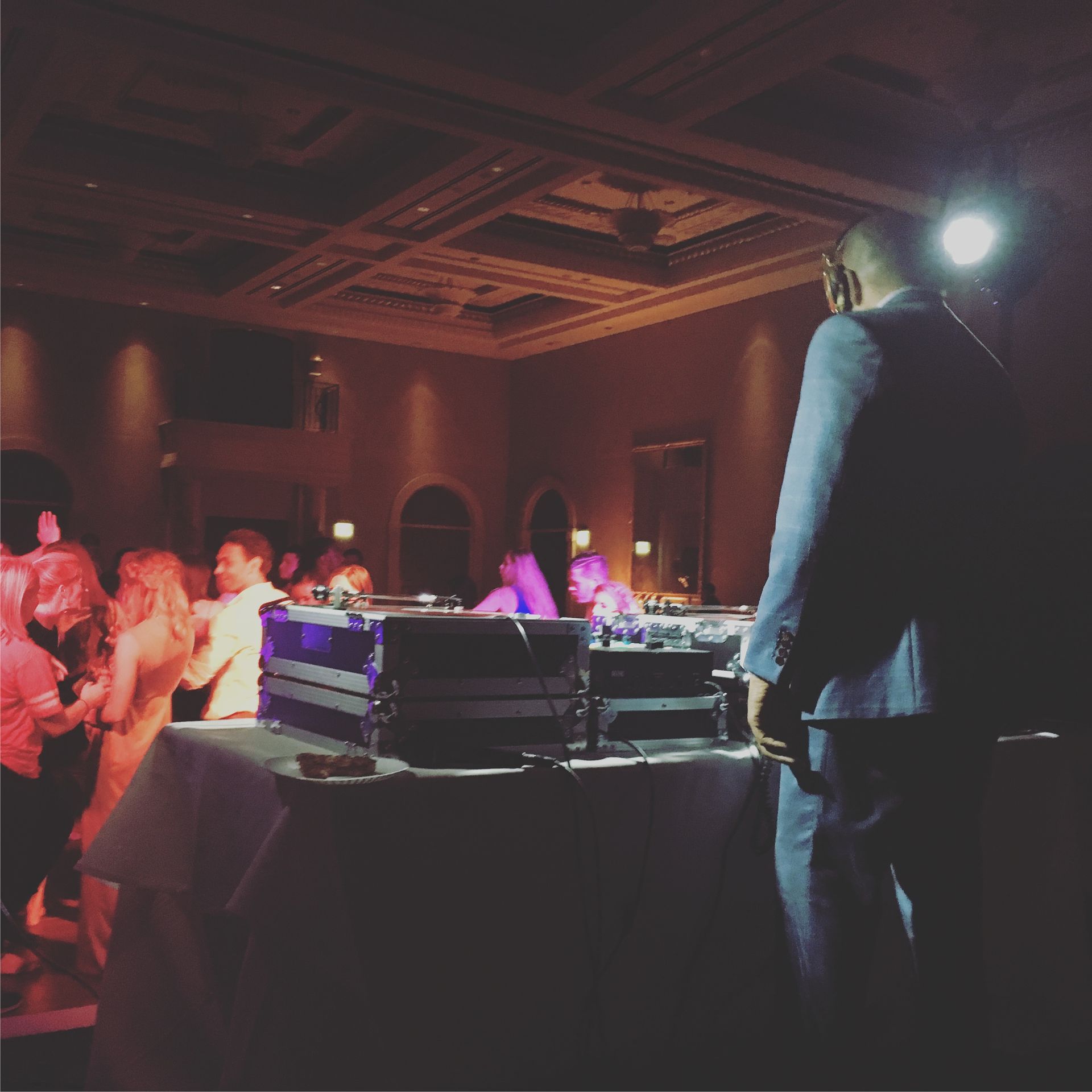 A DJ stands at a table with equipment, facing a dance floor in a dimly lit event space with guests dancing.