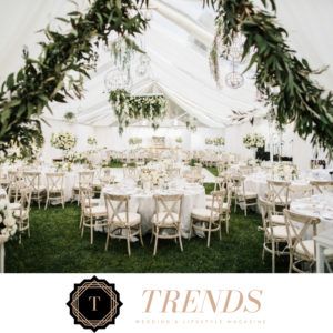 An elegant white wedding tent featuring round tables with cross-back chairs, adorned with lush greenery and hanging decor.
