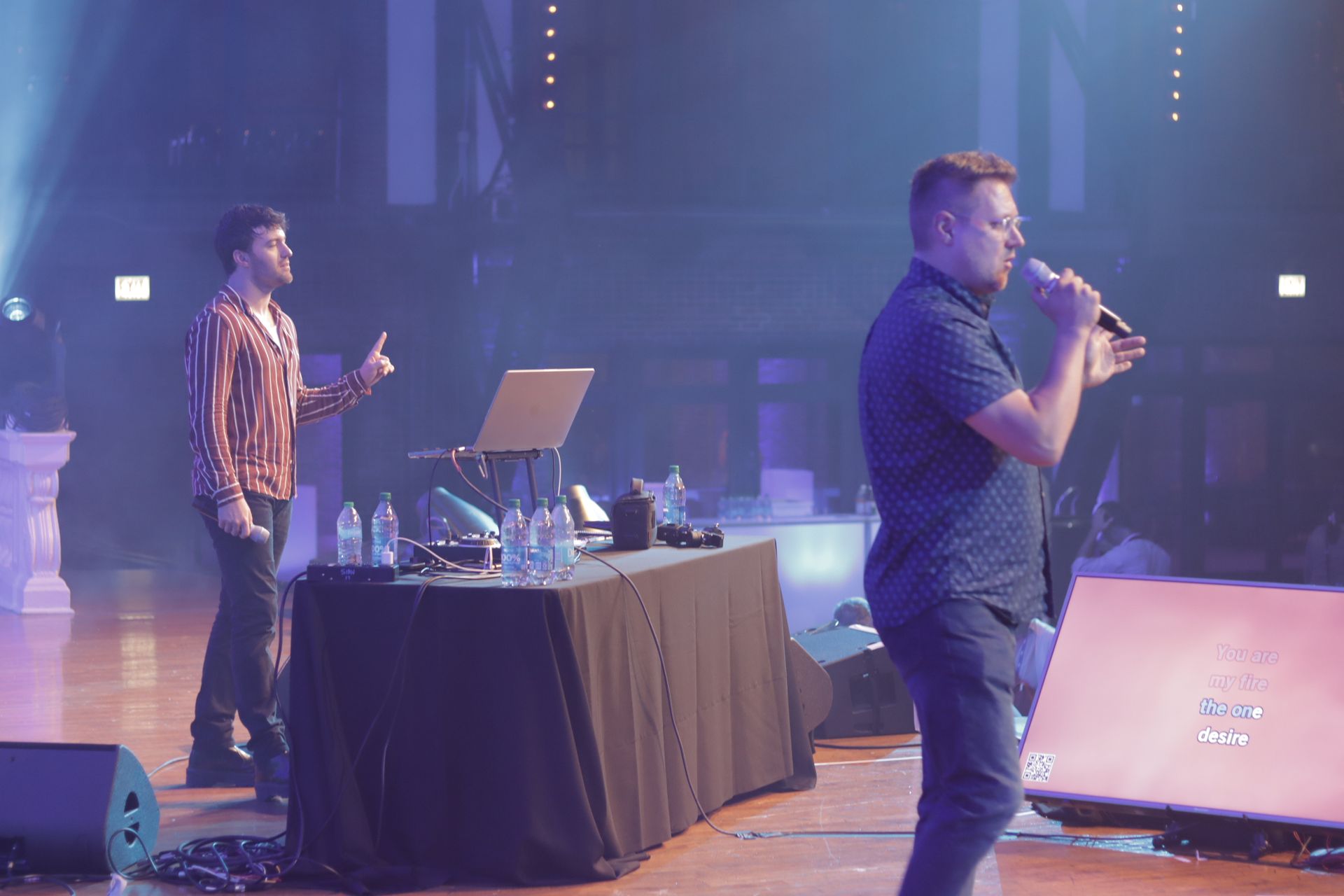 Two people perform on a stage with a table, laptop, and microphone in a dimly lit, professional event setting.
