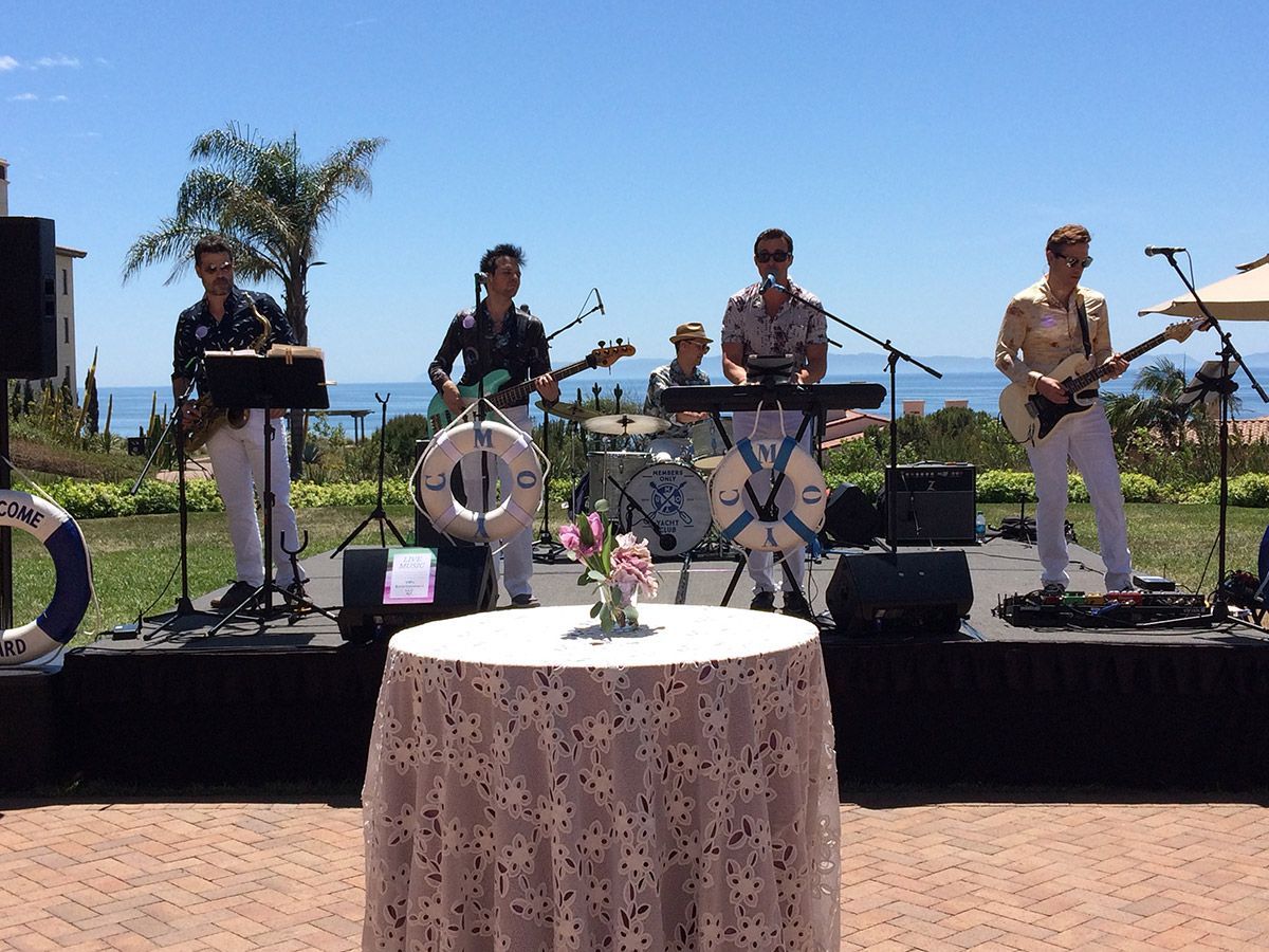 A live band performs on an outdoor stage overlooking the ocean, featuring a guitarist, bassist, drummer, and keyboardist.