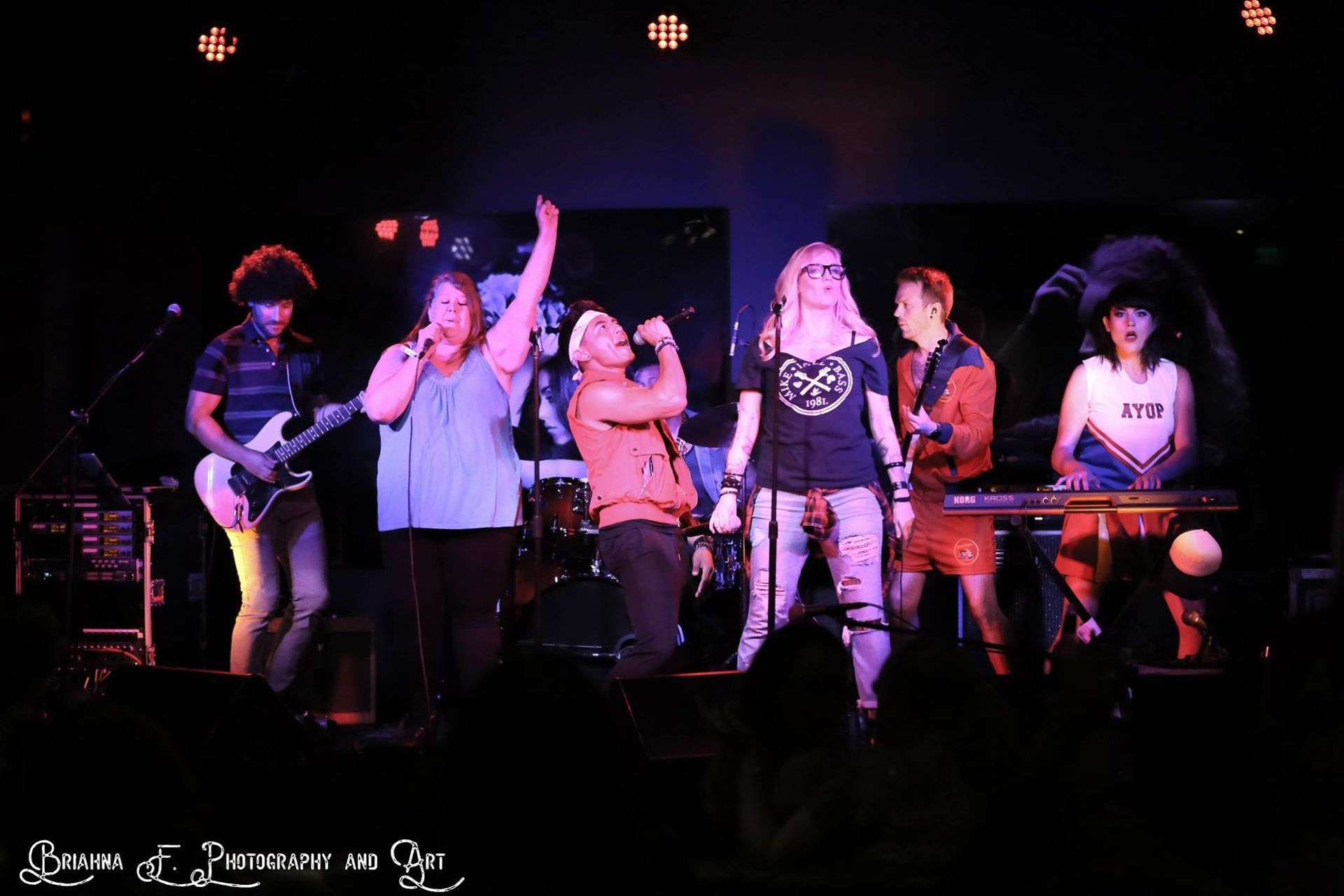 A band performs on a dim stage; a guitarist, two vocalists, a bassist, and a keyboardist are illuminated by stage lights.